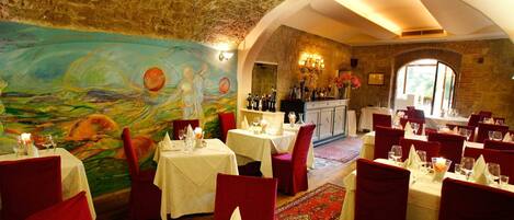 A restaurant with stone walls, red velvet chairs, and a mural on the wall. Tables are set with white tablecloths and candles.