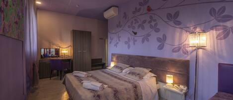 Un dormitorio con una cama grande, un escritorio y una silla. Se aprecia una pared con diseño floral y una lámpara sobre la mesita de noche.