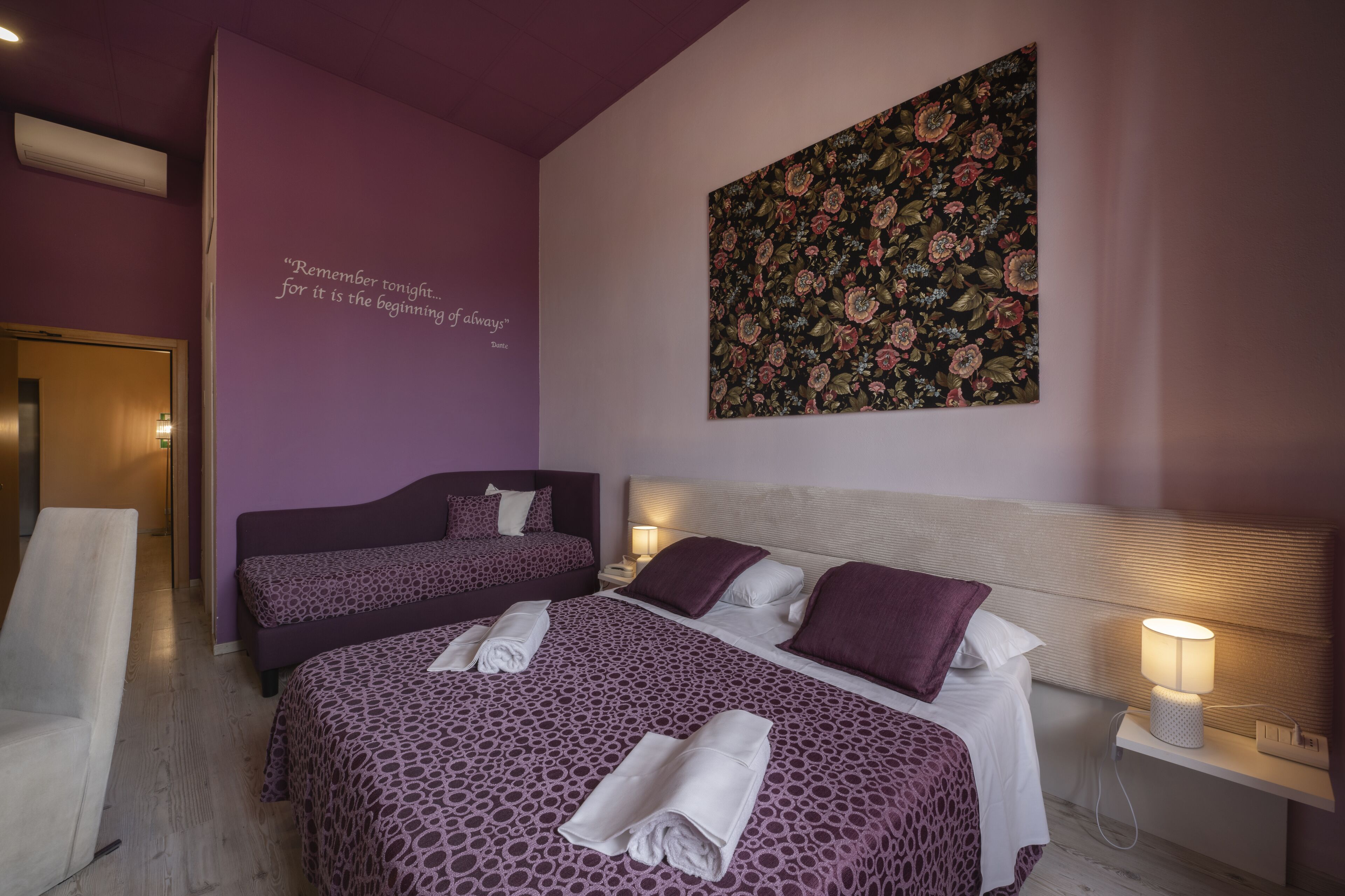 A hotel room with two beds, a purple wall with a quote, a floral painting, and a bedside table with a lamp.