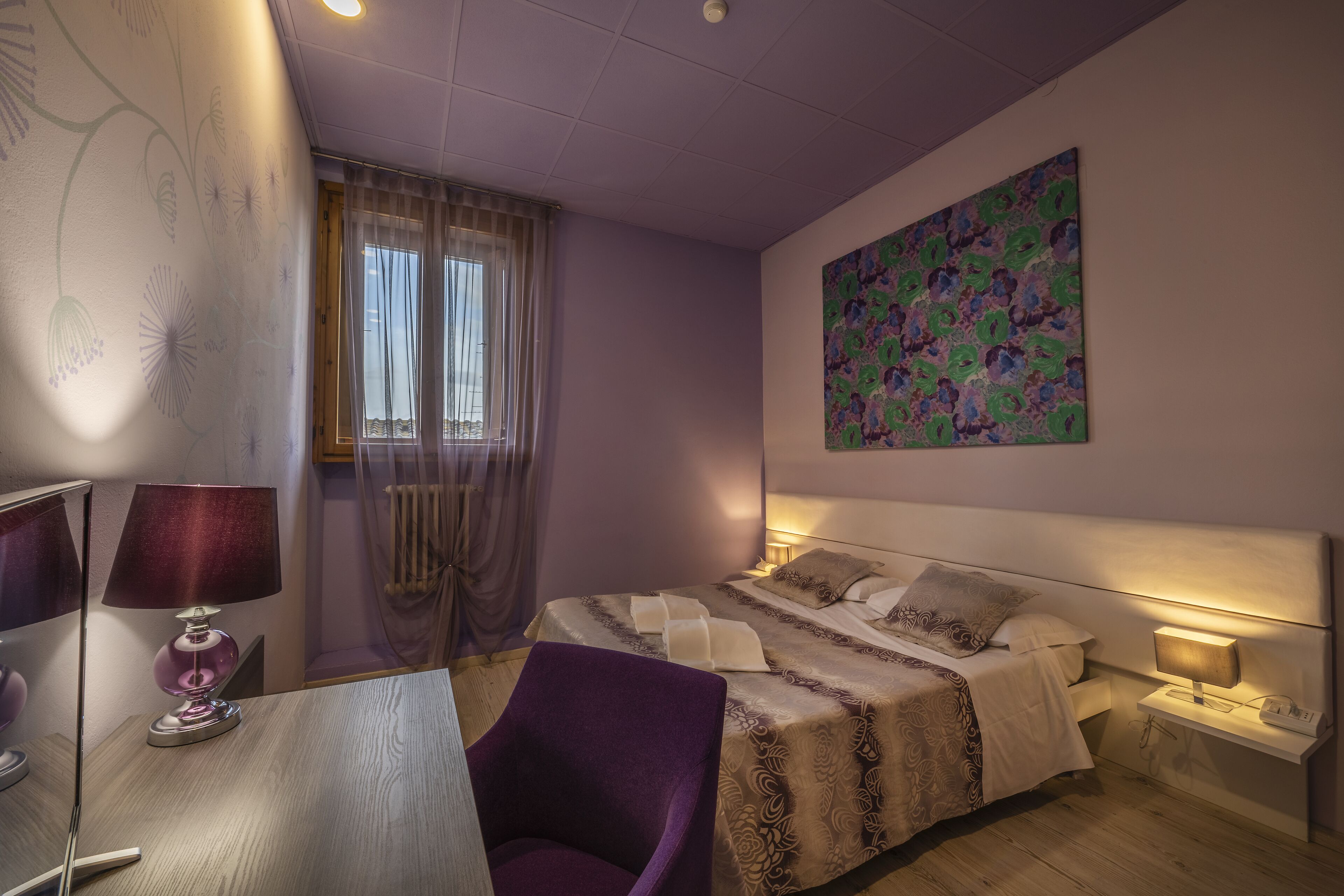A hotel room with a bed, bedside tables, a desk with a chair, a lamp, and a window with curtains.