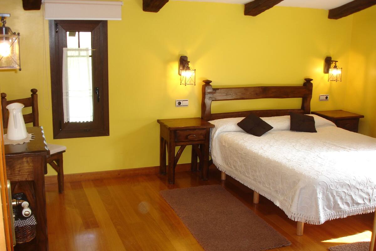 Un dormitorio con una cama de madera, dos mesitas de noche, una mesita auxiliar, una silla de madera, una pequeña mesa con una jarra y una ventana con persiana blanca.