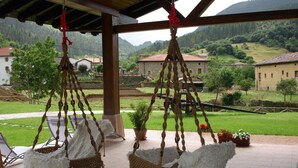 A covered outdoor seating area with wicker chairs and hanging decorations, overlooking a rural landscape with houses and greenery. - Lezamakoetxe (Sopuerta)