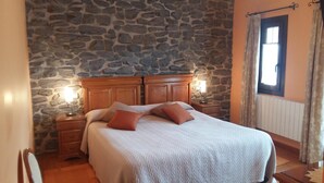 A bedroom with a stone wall, a wooden bed with two pillows, a nightstand with a lamp, and a window with curtains. - Lezamakoetxe (Sopuerta)