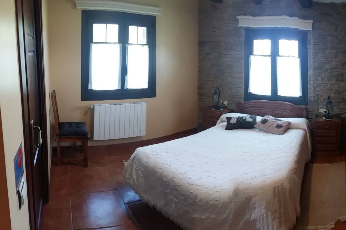 Un dormitorio con una cama, dos ventanas, un radiador, un armario de madera y una pequeña mesa con un jarrón.