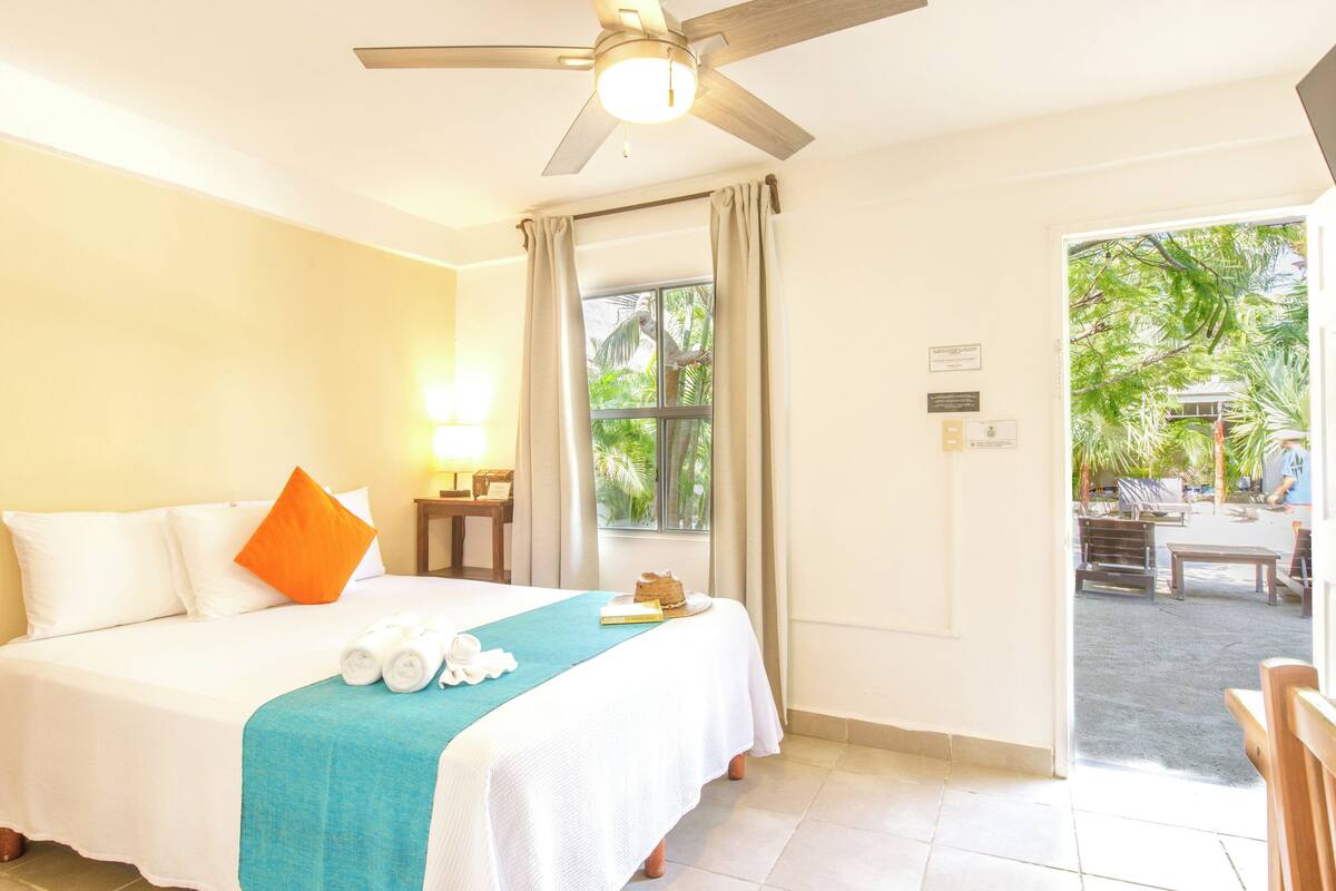 Une chambre d’hôtel avec un lit, un ventilateur de plafond, une fenêtre avec des rideaux, un bureau avec une lampe et une vue sur l’extérieur.