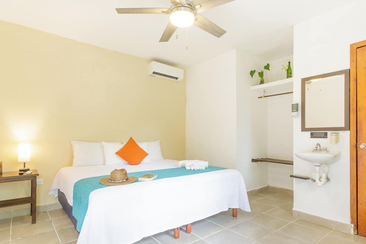 Une chambre d’hôtel comprenant un lit, un ventilateur de plafond, un appareil de climatisation, une table de chevet avec une lampe, une salle de bain avec un lavabo et un miroir, et une porte en bois.