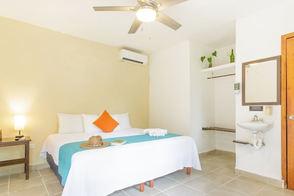 Habitación de hotel con cama, ventilador de techo, aire acondicionado, mesita de noche con lámpara, baño con lavamanos y espejo, y puerta de madera.