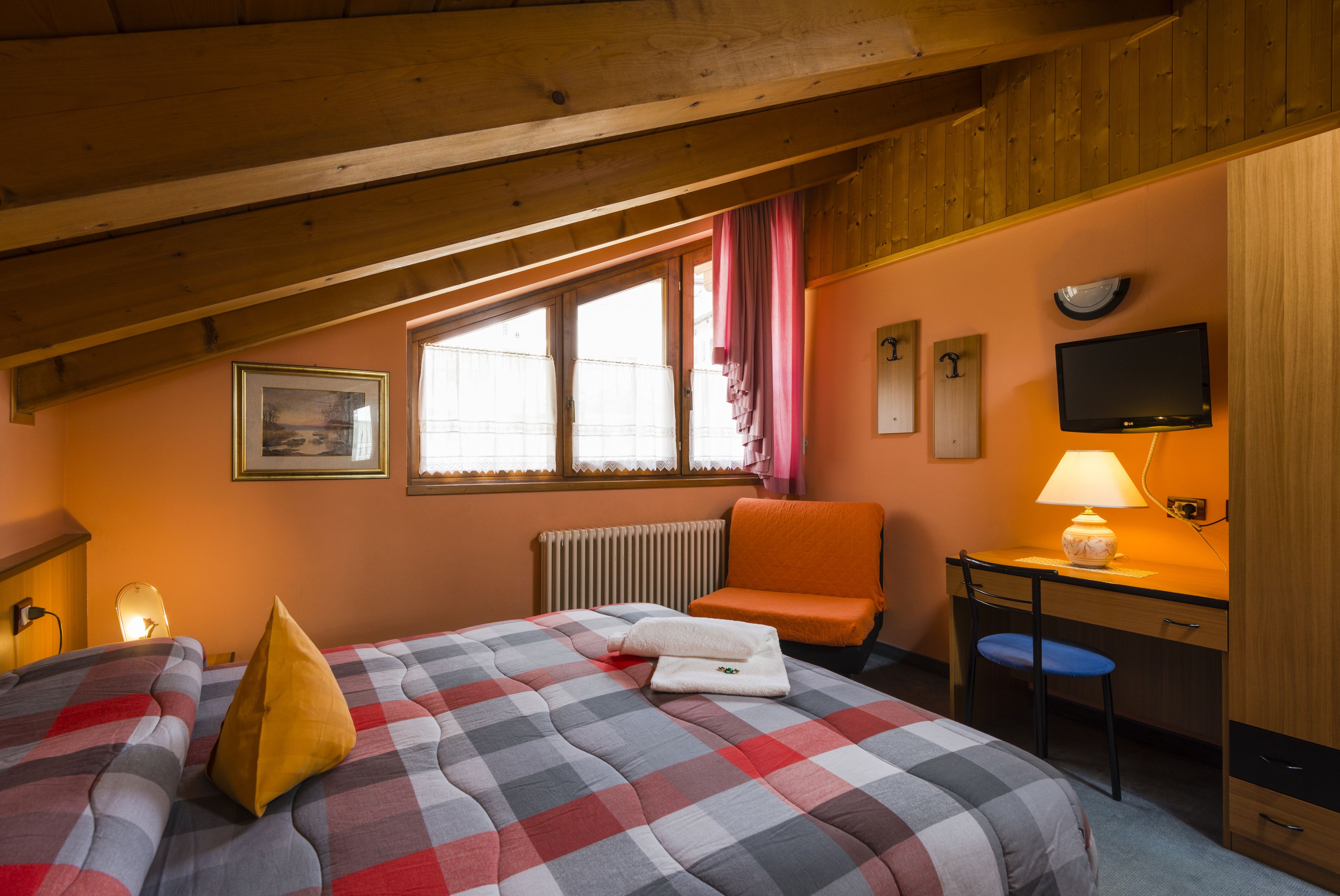 A bedroom with a checkered bedspread, a desk with a lamp, a chair, a television, and a window with curtains.