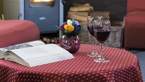 Una habitación acogedora con una estufa de leña, una mesa con mantel rojo de lunares, dos copas de vino y un jarrón con flores.