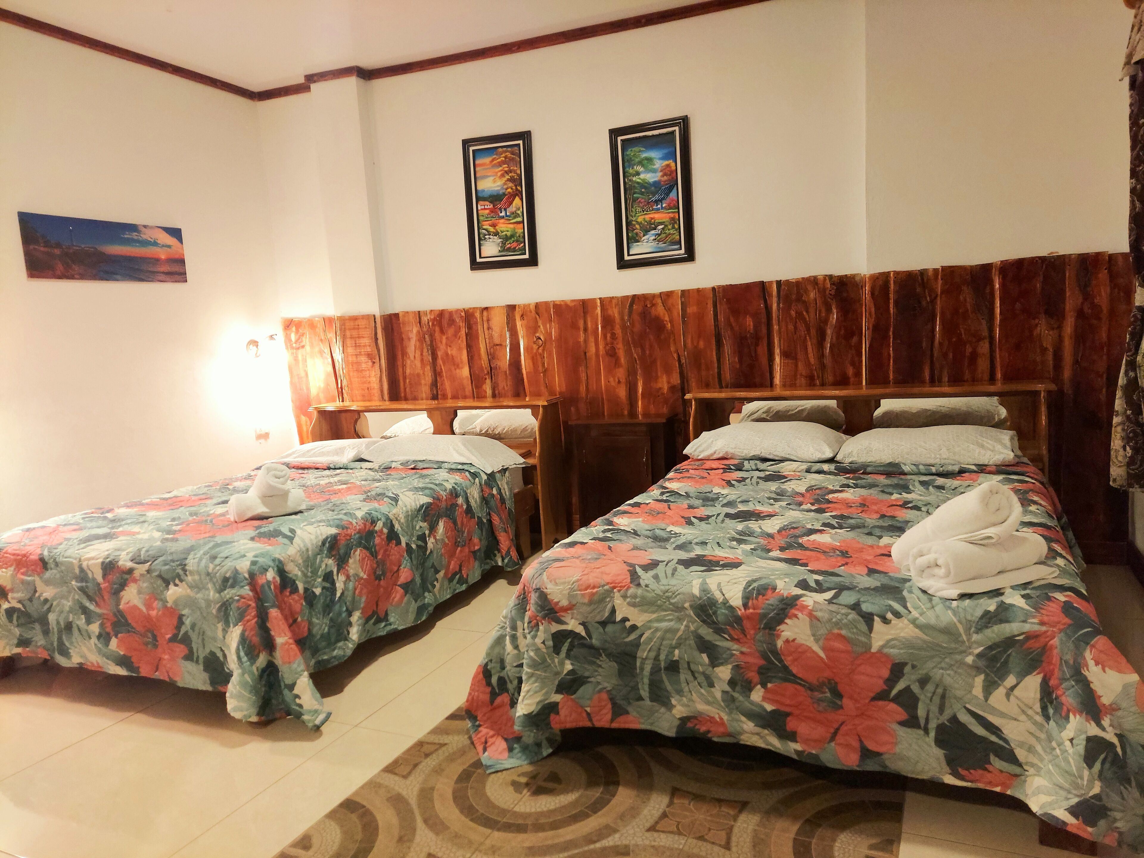 A hotel room with two single beds, wooden headboards, floral bedspreads, and two framed pictures on the wall.