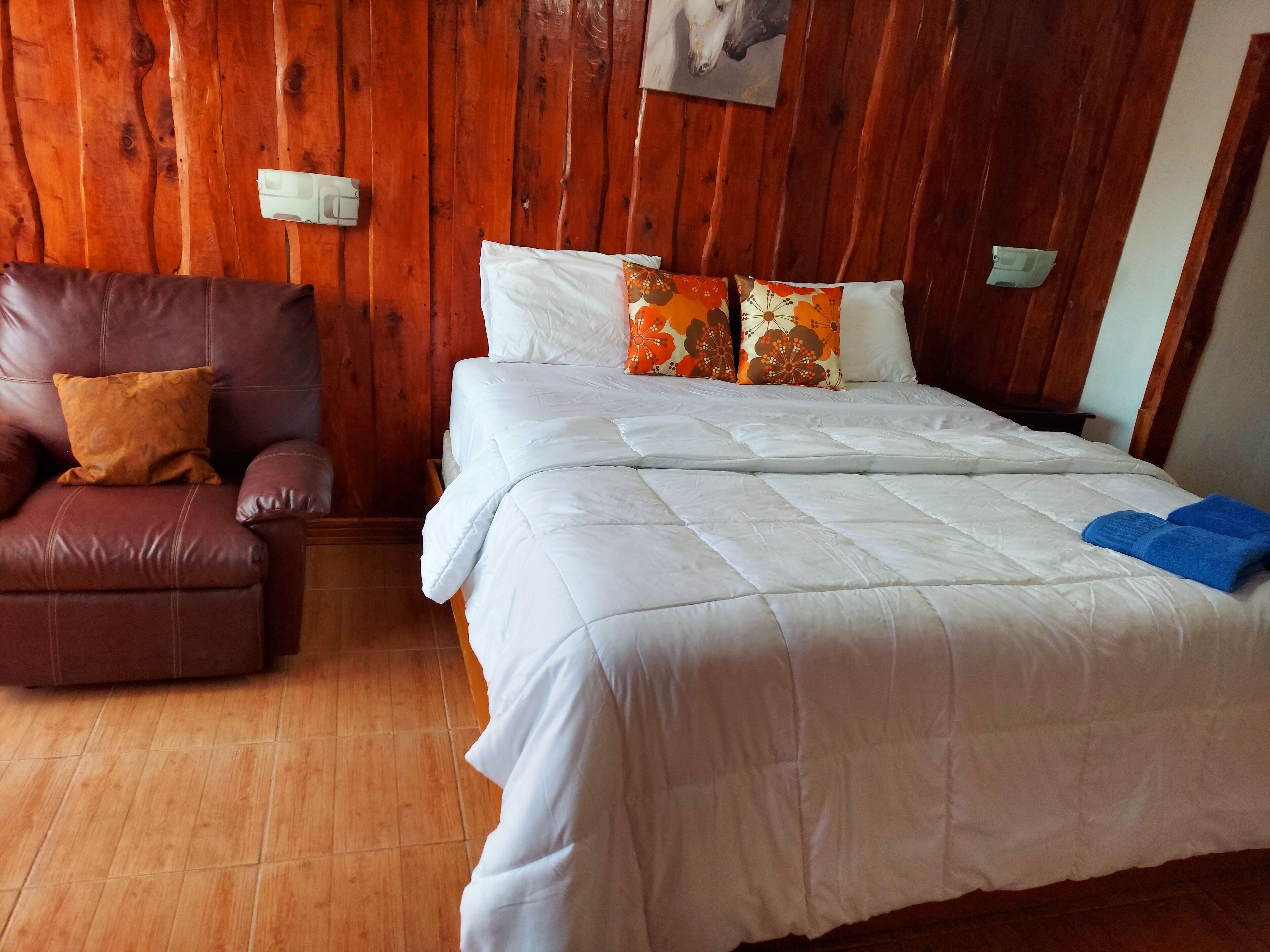 A room with a wooden paneled wall, a brown leather chair, a bed with white bedding and orange floral pillows, and a blue towel on the bed.