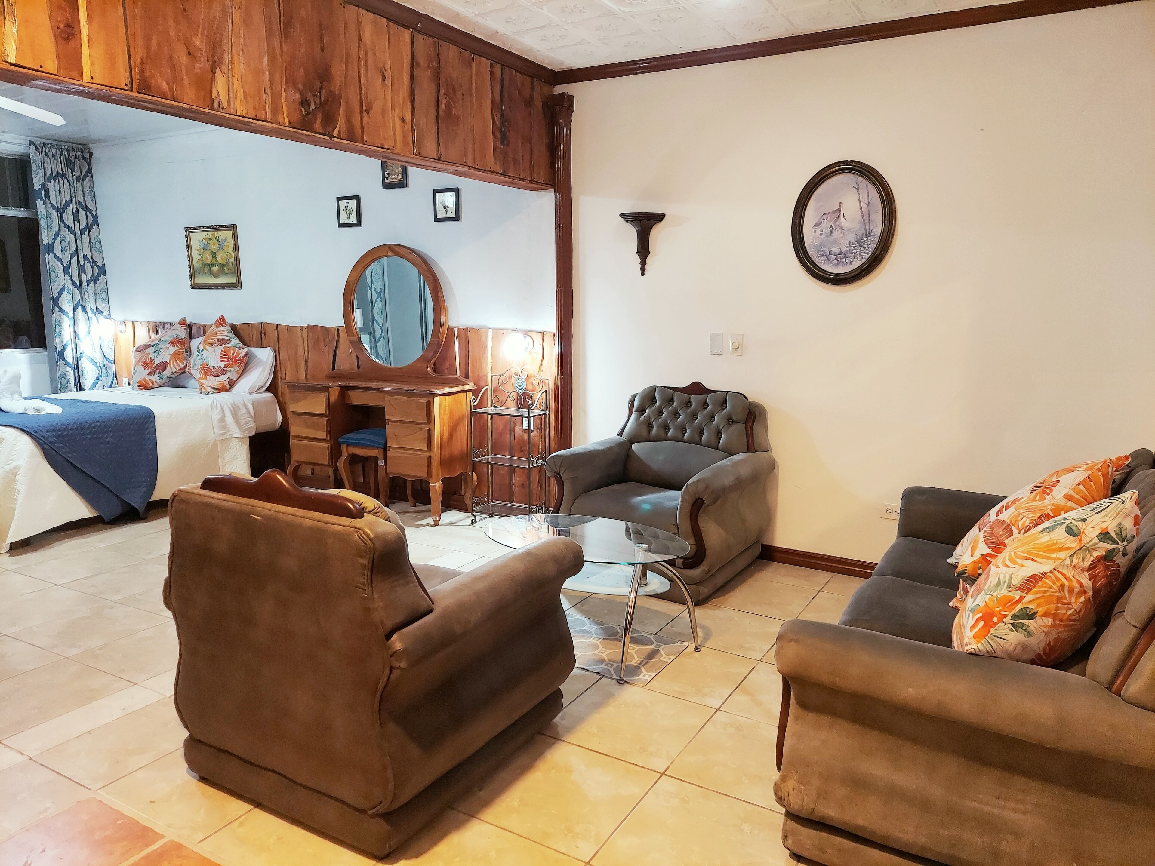 A living room with a sofa, armchair, and a small table. There is a bed with a blue blanket and a wooden headboard. A mirror hangs on the wall, and there are framed pictures above it.