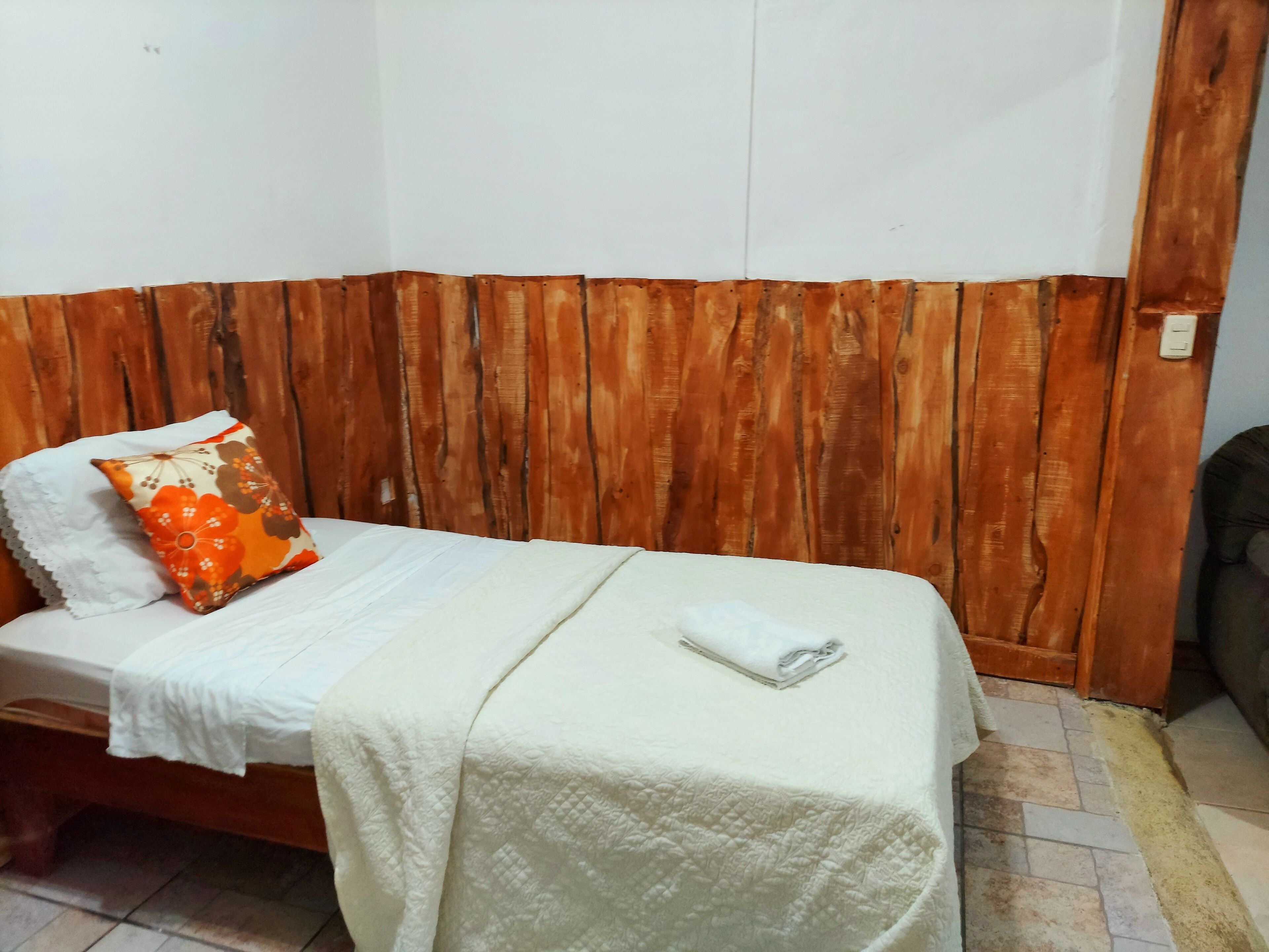 Un lit simple recouvert d’une courtepointe blanche, avec un oreiller blanc et un oreiller à motifs floraux orange. Les murs de la pièce sont recouverts de lambris en bois et le sol est carrelé.