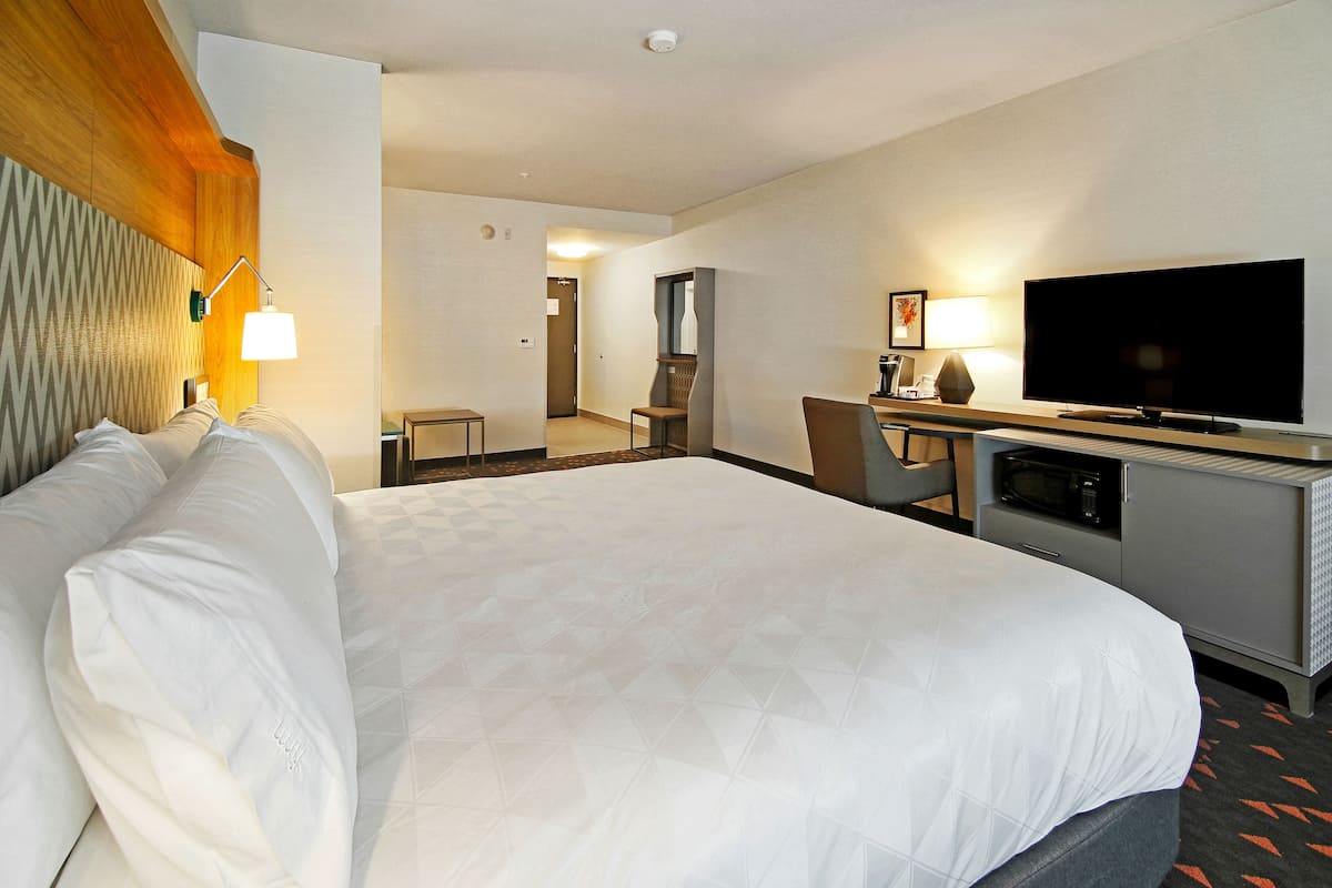 A hotel room with a large bed, a desk with a flat-screen TV, a microwave, and a seating area with a chair and a table.
