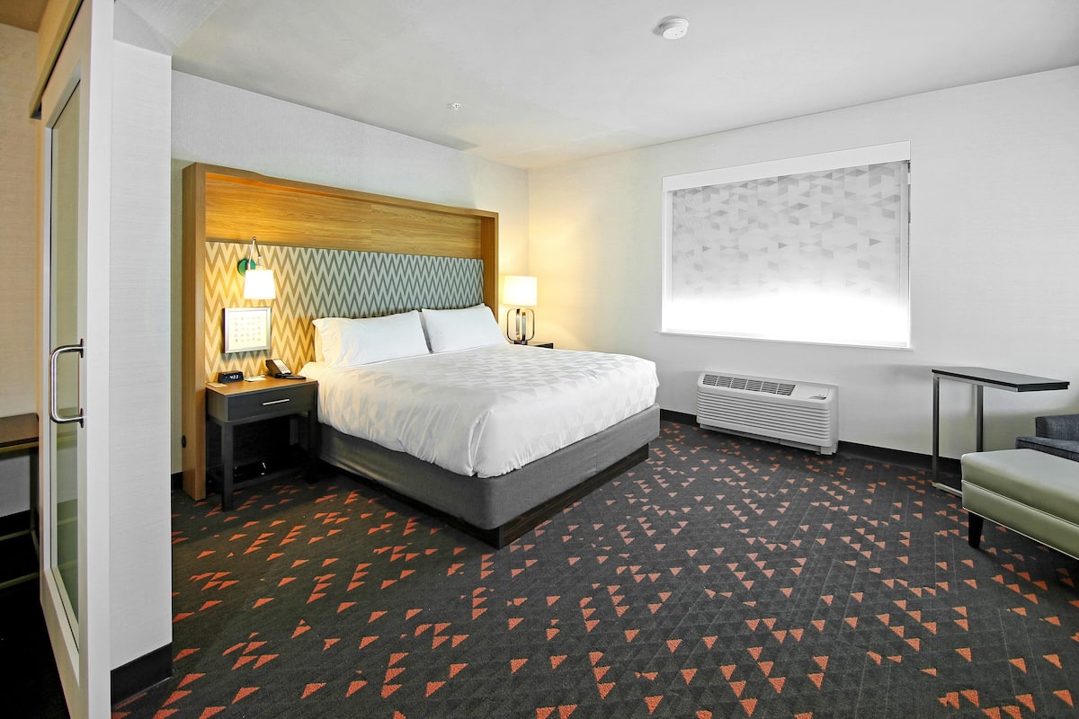 A hotel room with a bed, bedside tables, a nightstand, a window with blinds, a chair, and a carpet with a geometric pattern.