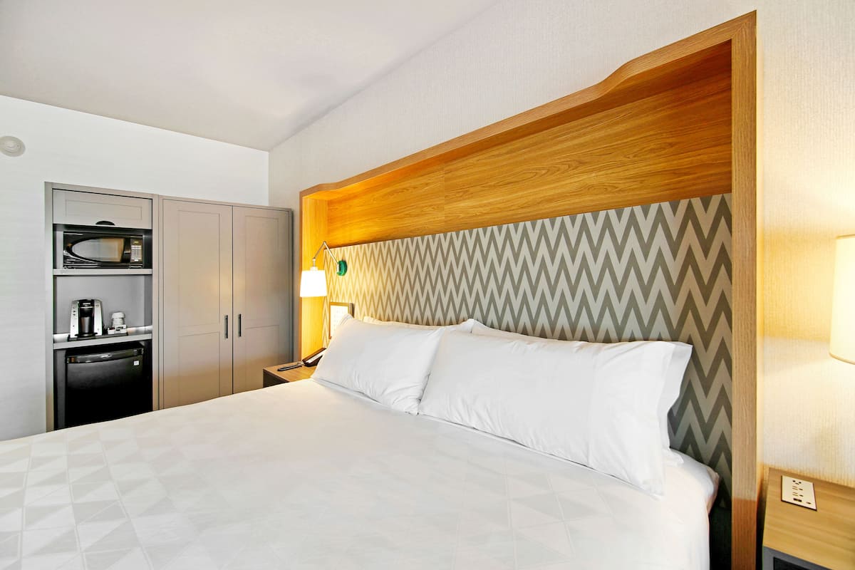 A hotel room with a bed, a built-in microwave, a coffee machine, and a small desk with a lamp.