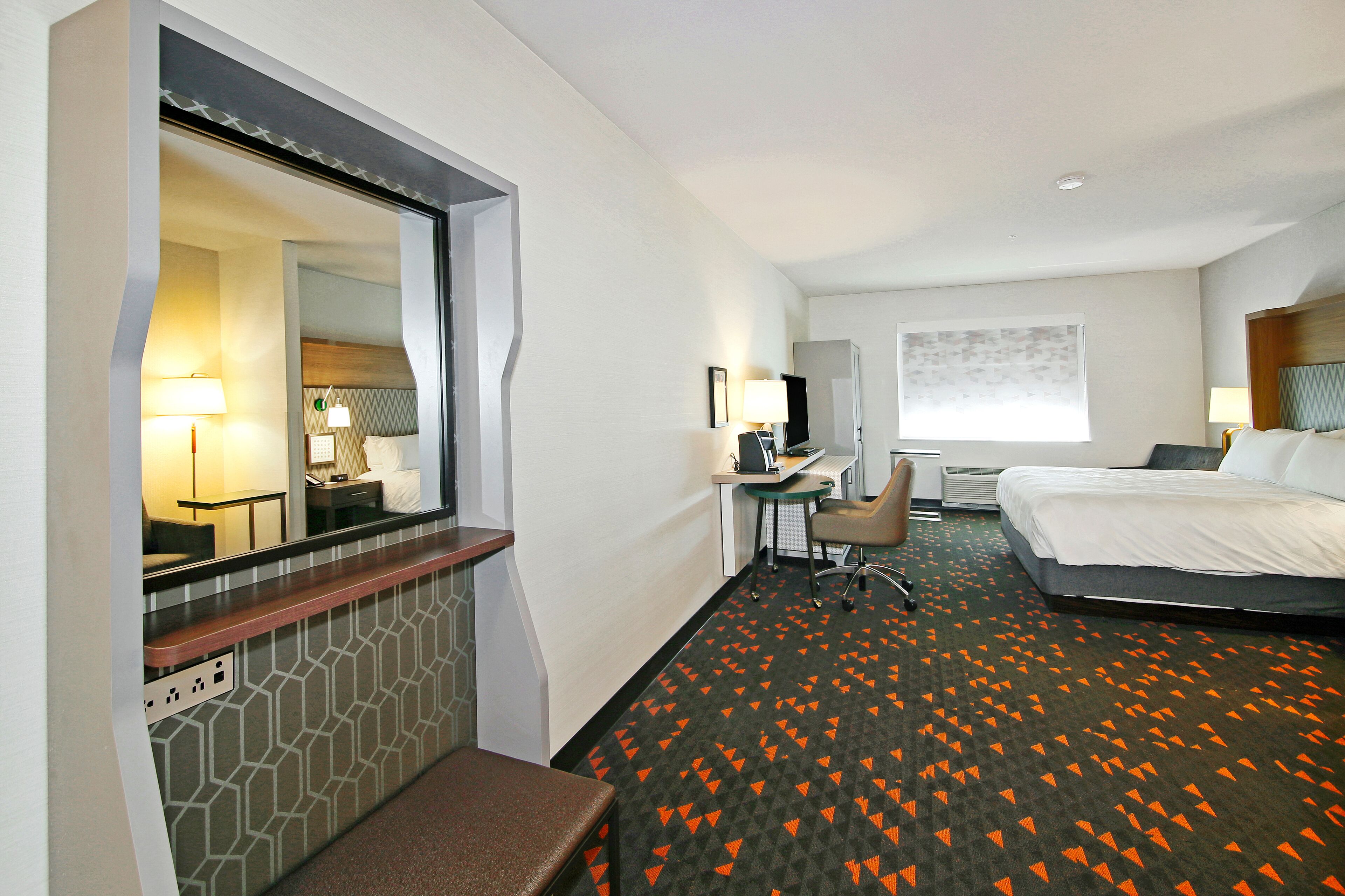 A hotel room with a bed, desk, chair, and a large mirror.