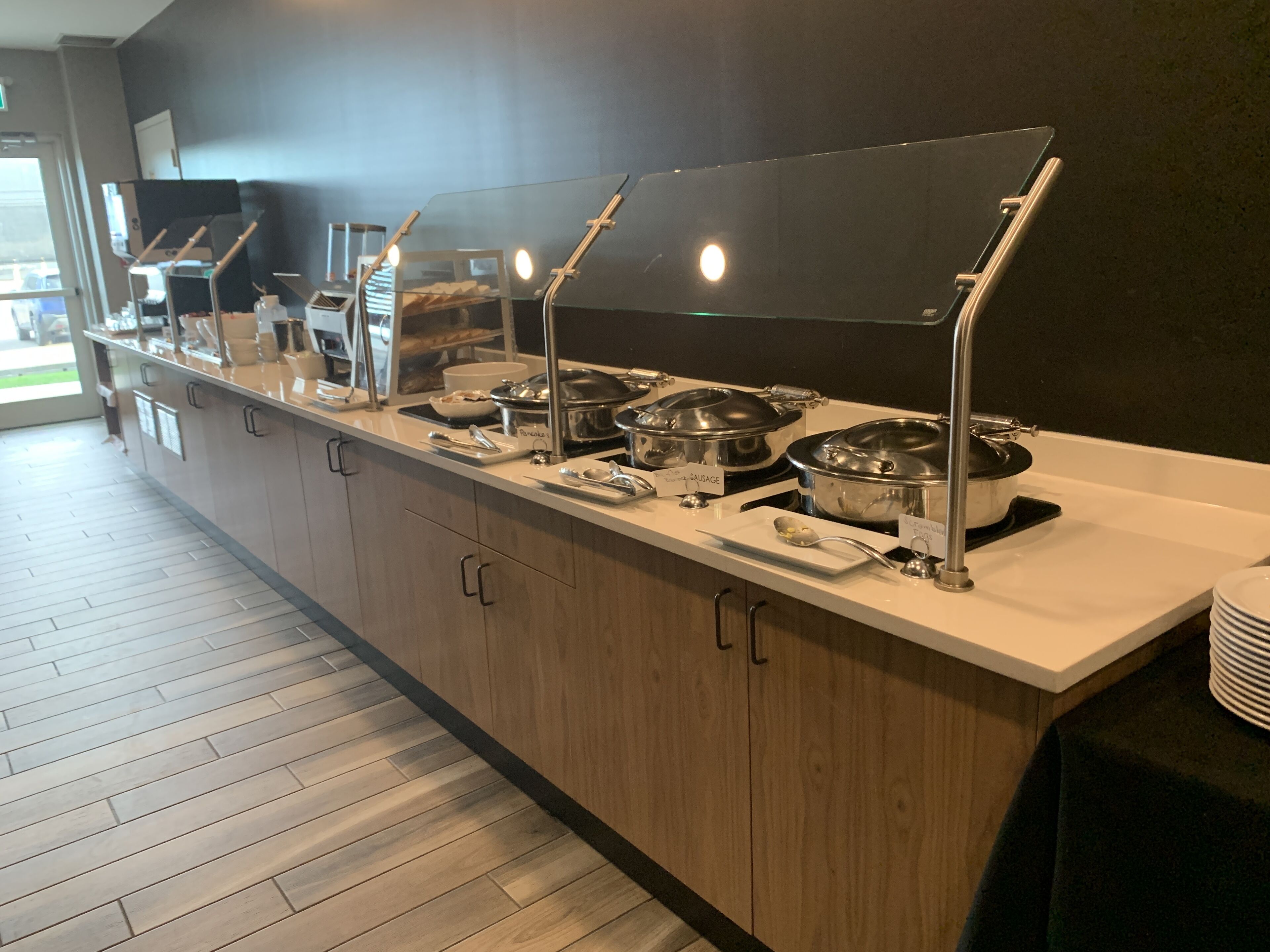 Buffet montado com várias estações de serviço, cada uma com um prato coberto e uma placa indicando o tipo de comida.