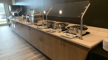 Buffet montado com várias estações de serviço, cada uma com um prato coberto e uma placa indicando o tipo de comida.