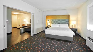 Habitación de hotel con una cama grande, una mesita de noche con lámpara, una silla y una pequeña mesa. Se ve un pasillo que conduce a otra habitación.