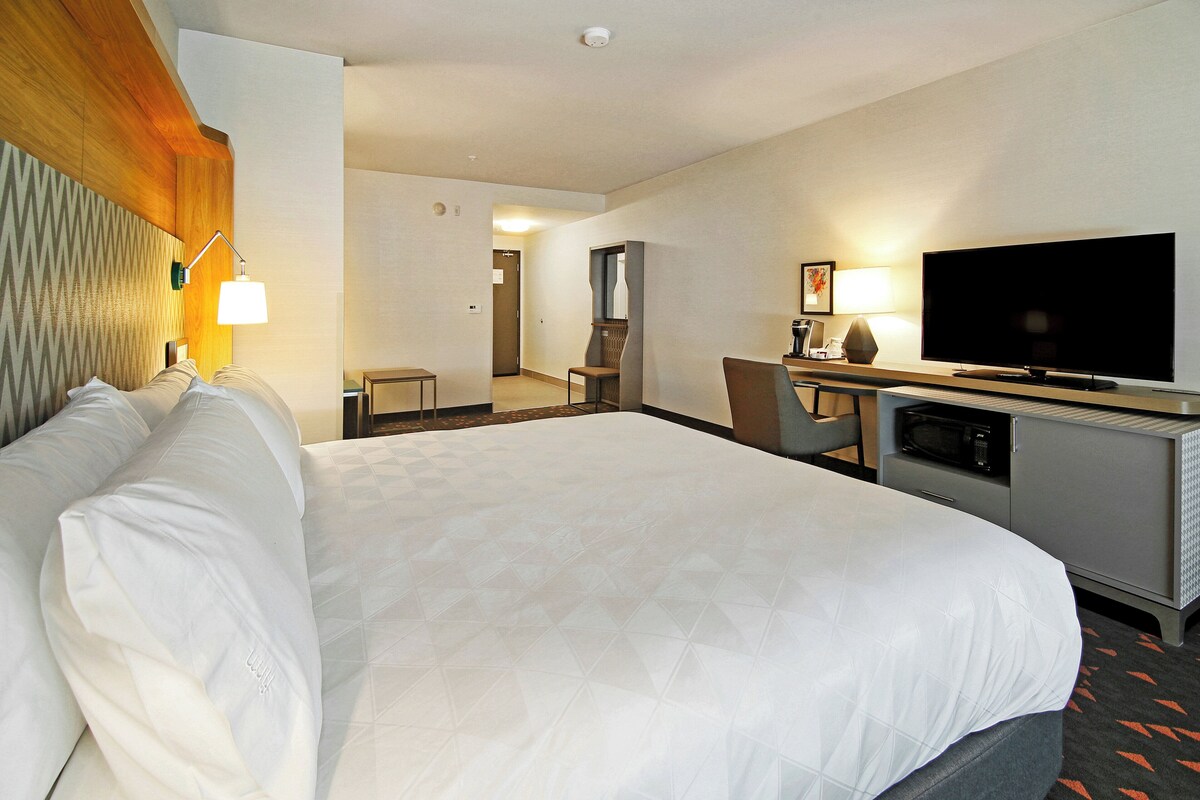 A hotel room with a large bed, a desk with a flat-screen TV, a microwave, and a seating area with a chair and table.