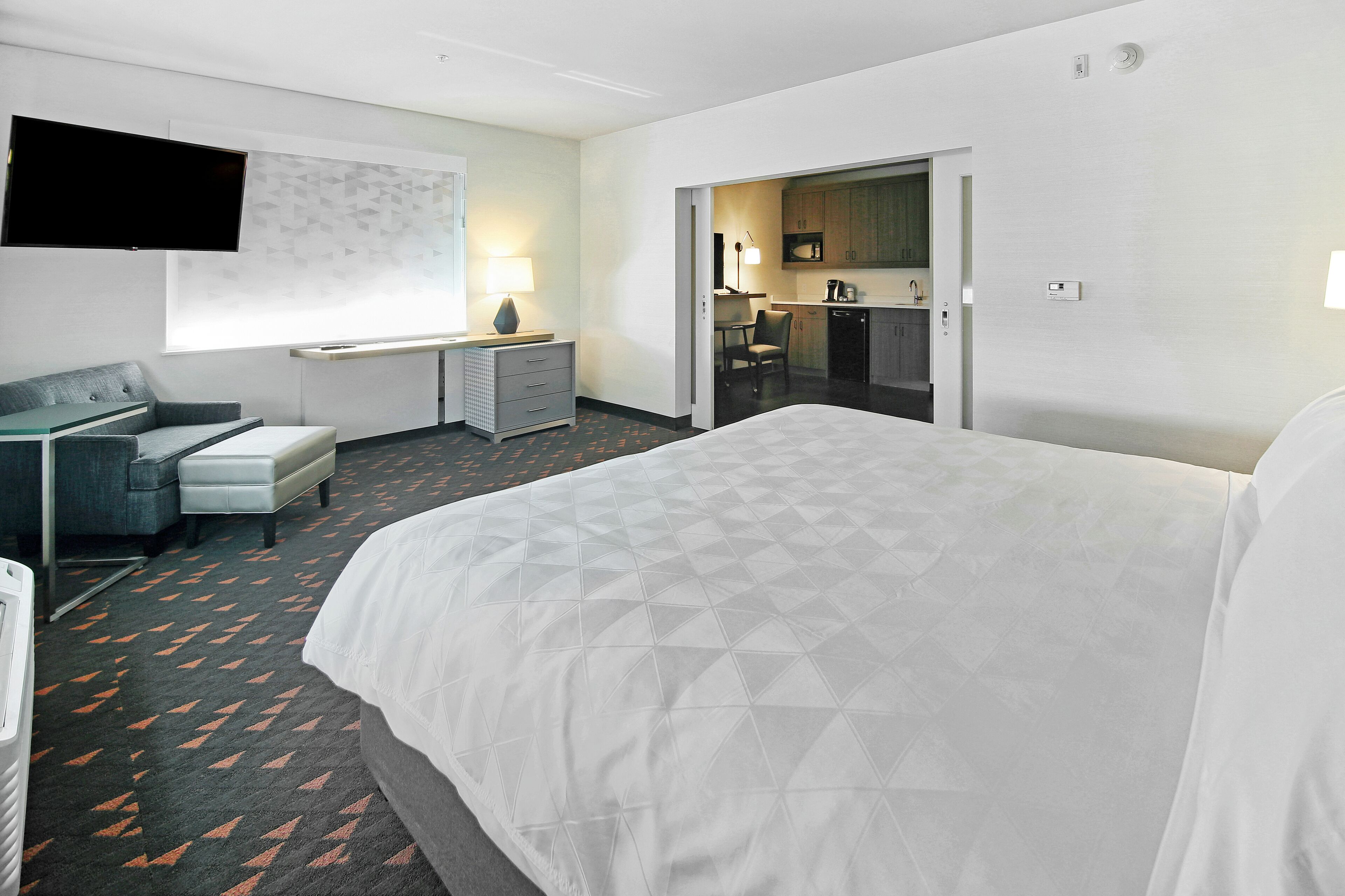 A hotel room with a large bed, a desk with a chair, a TV mounted on the wall, and a kitchenette in the adjacent room.