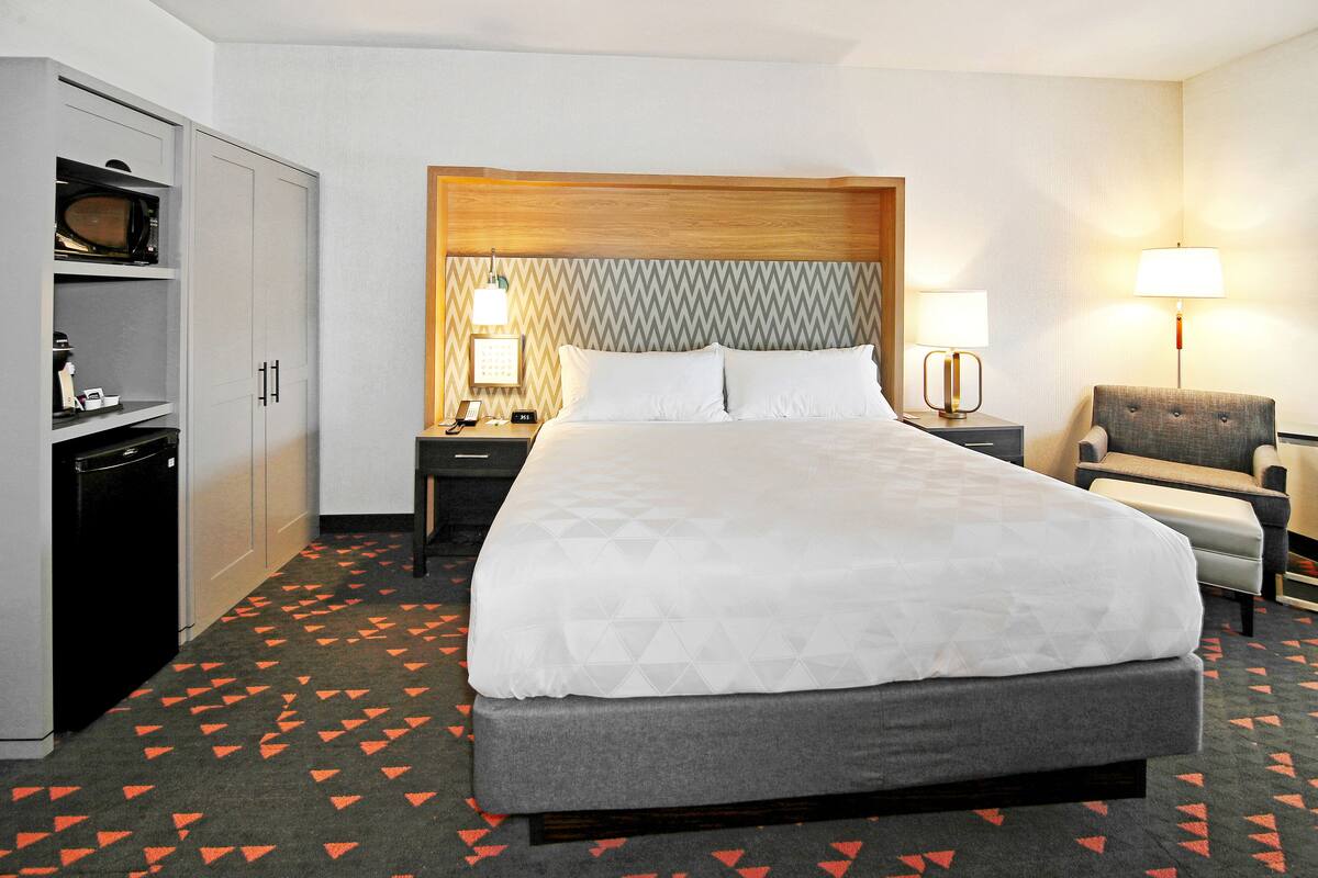 A hotel room with a large bed, a desk with a lamp, a chair, and a wardrobe.