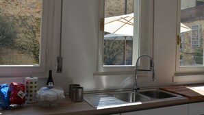 A kitchen with a stainless steel sink, a window with a white umbrella, and a view of a stone building. - Dimora 16 Rooms & Garden (Prato)