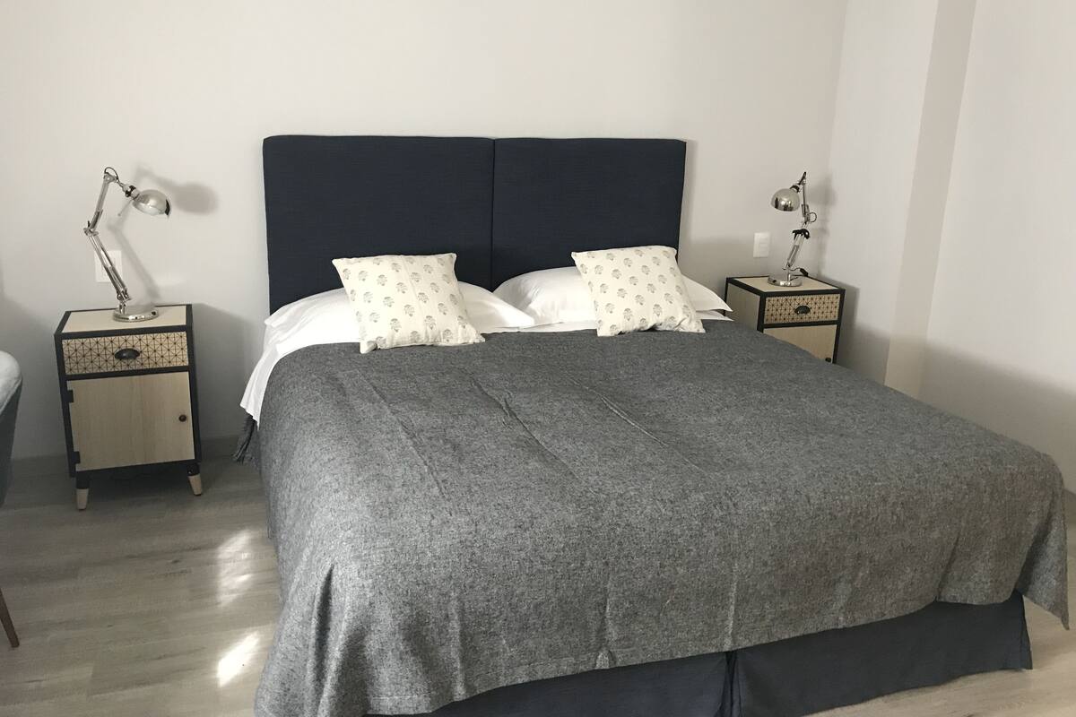 A neatly made bed with a grey blanket, two bedside tables with lamps, and a plain white wall.