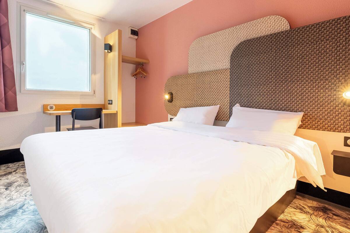 Een net opgemaakt bed met witte lakens, een groot raam met gordijnen, een klein bureau met stoel en een wandlamp.