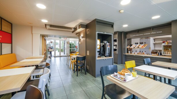 Un café moderne doté de plusieurs espaces de détente, comprenant des tables et des chaises, ainsi qu’un comptoir où l’on peut commander des plats et des boissons.