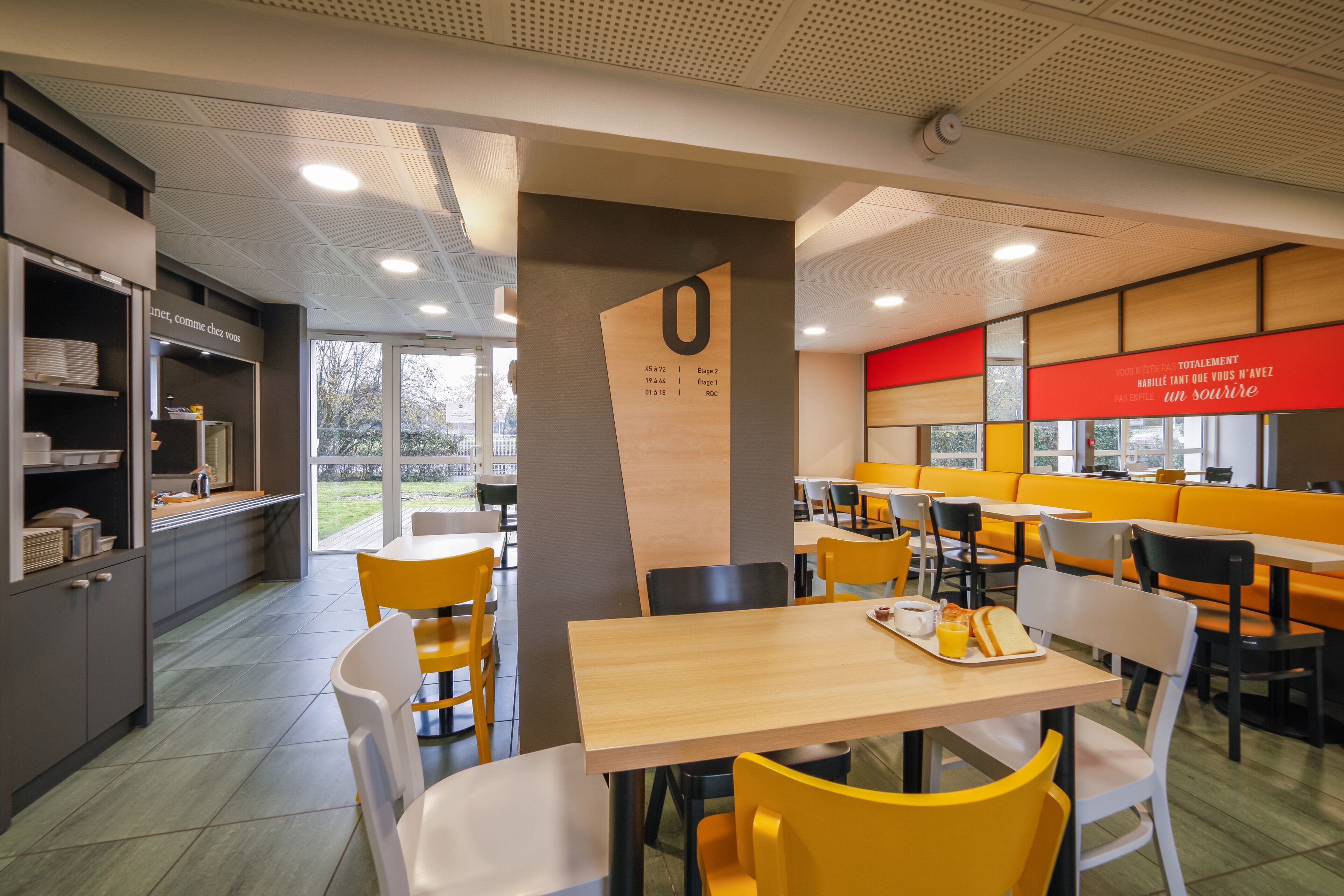 Eine moderne Cafeteria mit Holztischen und gelben Stühlen, ein Küchenbereich mit Regalen sowie ein großes rotes Schild an der Wand.