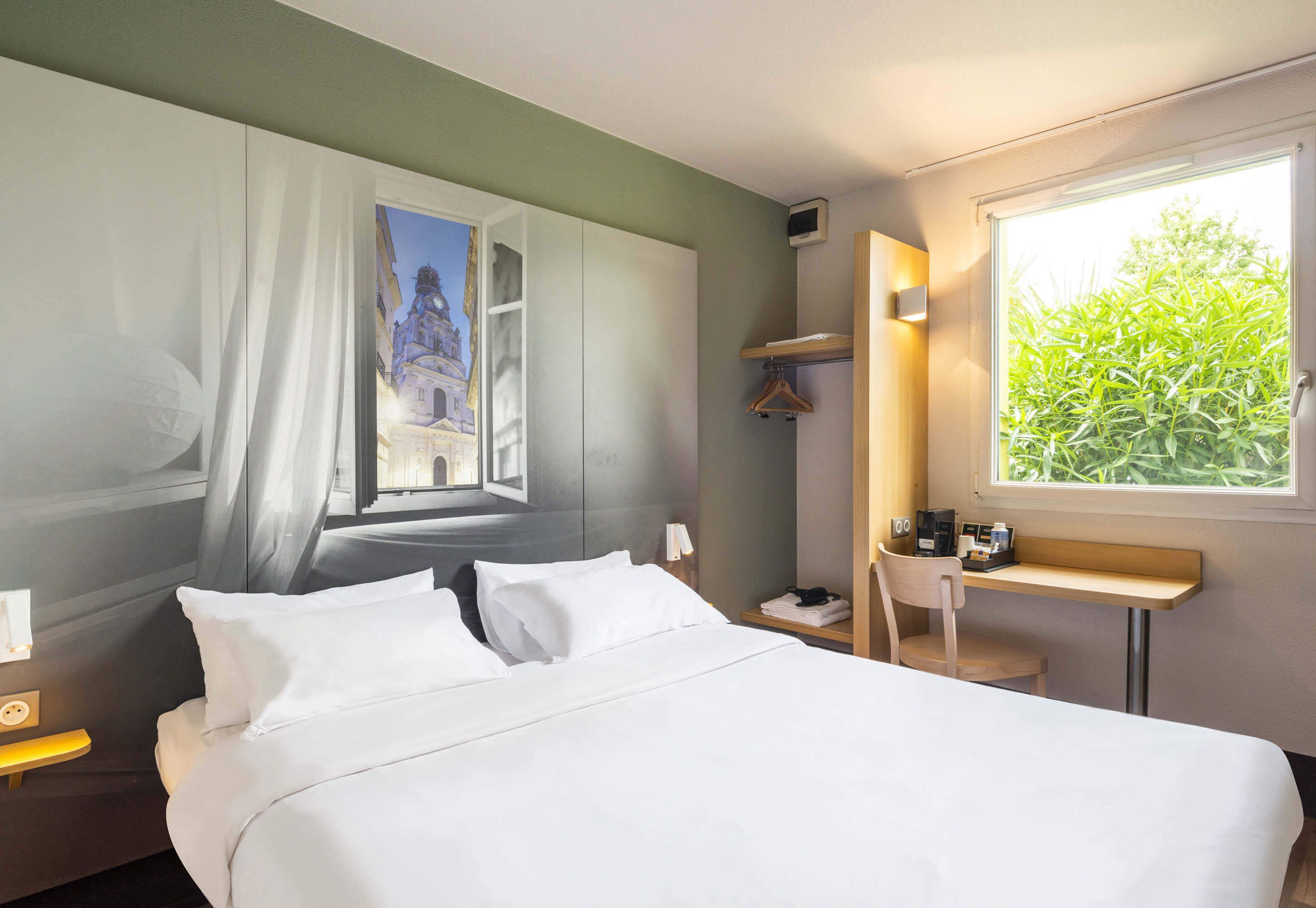 Une chambre d’hôtel comprenant un lit, une table de chevet, un bureau, une chaise, une fenêtre donnant sur un espace verdoyant et un tableau accroché au mur.