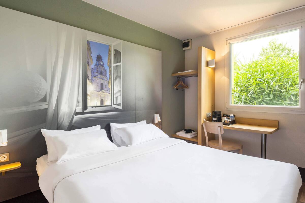 Hotelkamer met een bed, nachtkastje, bureau, stoel, raam met uitzicht op groen, en een schilderij aan de muur.