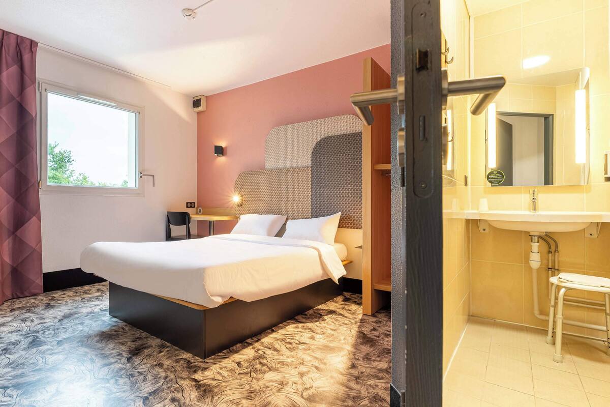 Een hotelkamer met een groot bed, een nachtkastje, een stoel, een badkamer met een wastafel en spiegel, en een raam met gordijnen.