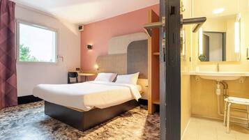 Habitación de hotel con una cama grande, una mesita de noche, una silla, baño con lavamanos y espejo, y ventana con cortinas.