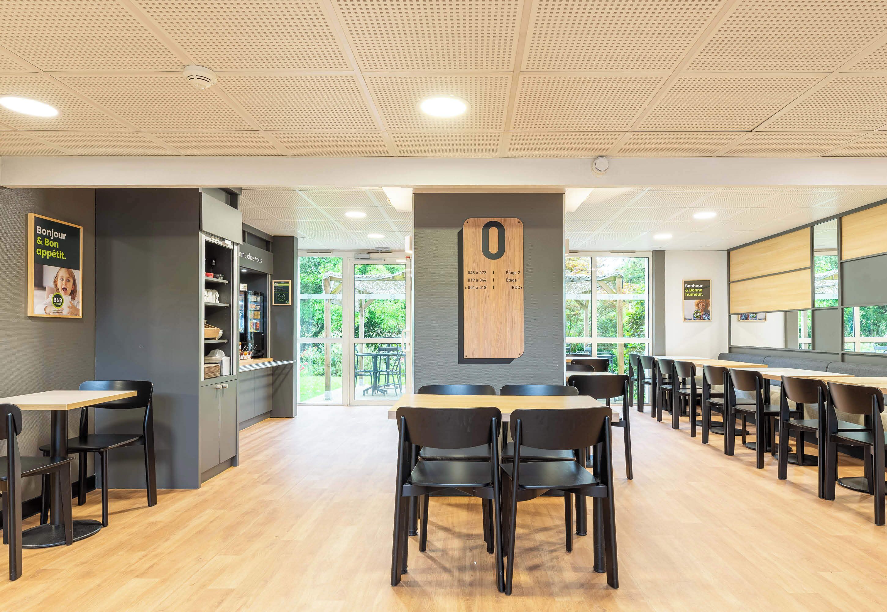 A modern dining area with wooden tables and chairs, a large wooden number '0' sign, and a view of greenery outside.