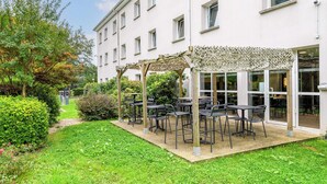 Un espace extérieur aménagé avec des tables et des chaises, entouré de verdure et bordé d’un b âtiment doté de grandes fenêtres.