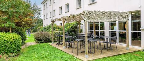 Un espace extérieur aménagé avec des tables et des chaises, entouré de verdure et bordé d’un bâtiment doté de grandes fenêtres.
