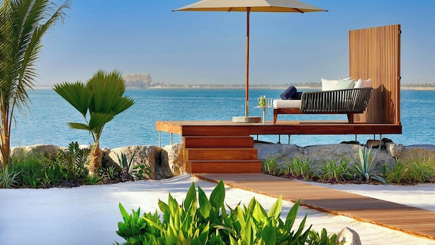 A modern outdoor seating area with a wooden deck, a cushioned bench, and a large umbrella. - The Ritz-Carlton Ras Al Khaimah, Al Hamra Beach (Ras Al Khaimah)