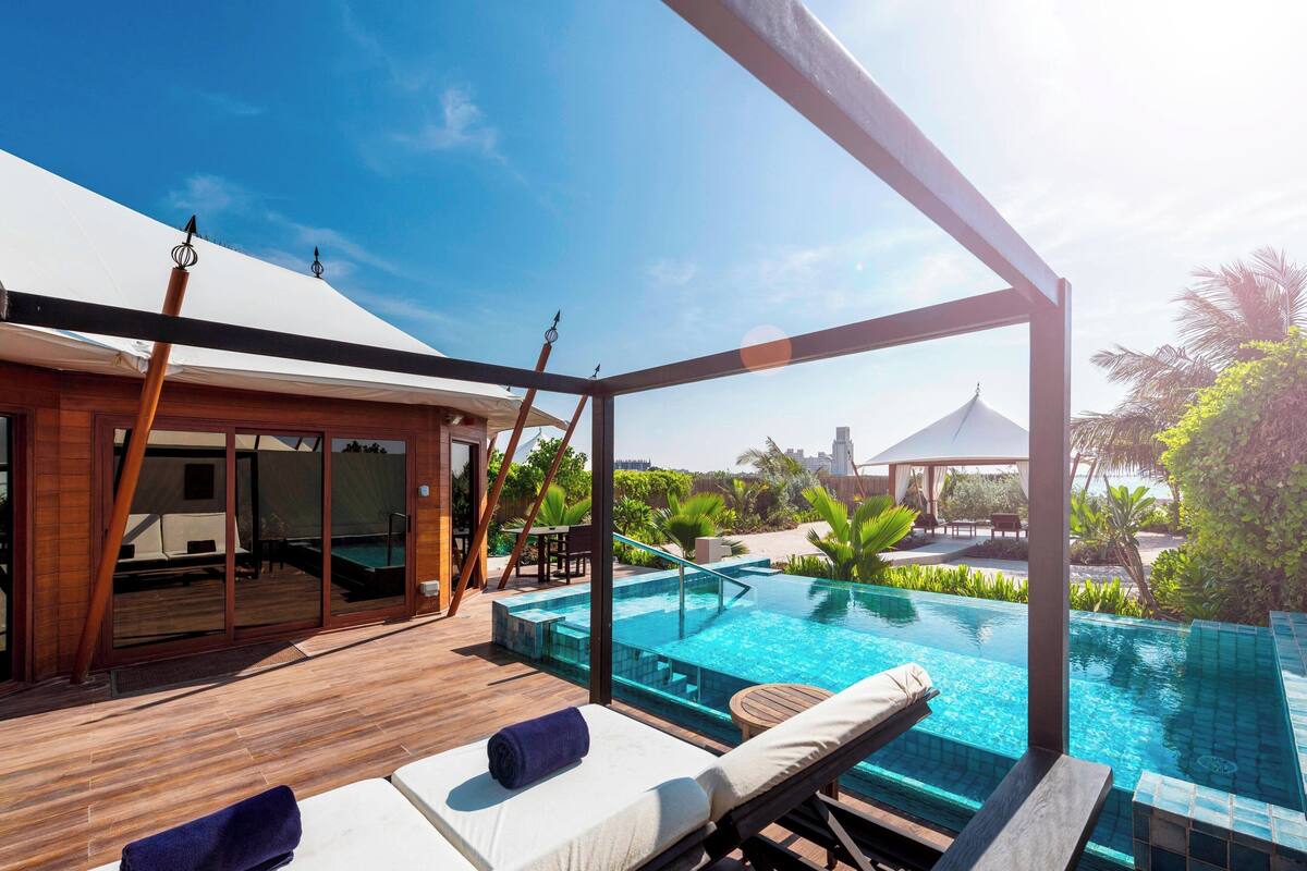 A poolside area with lounge chairs, a wooden deck, and a glass-enclosed room with a view of the pool and surrounding greenery.