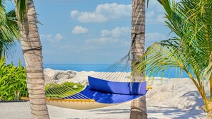 A hammock on a sandy beach with palm trees, a clear sky, and the ocean in the background. - The Ritz-Carlton Ras Al Khaimah, Al Hamra Beach (Ras Al Khaimah)