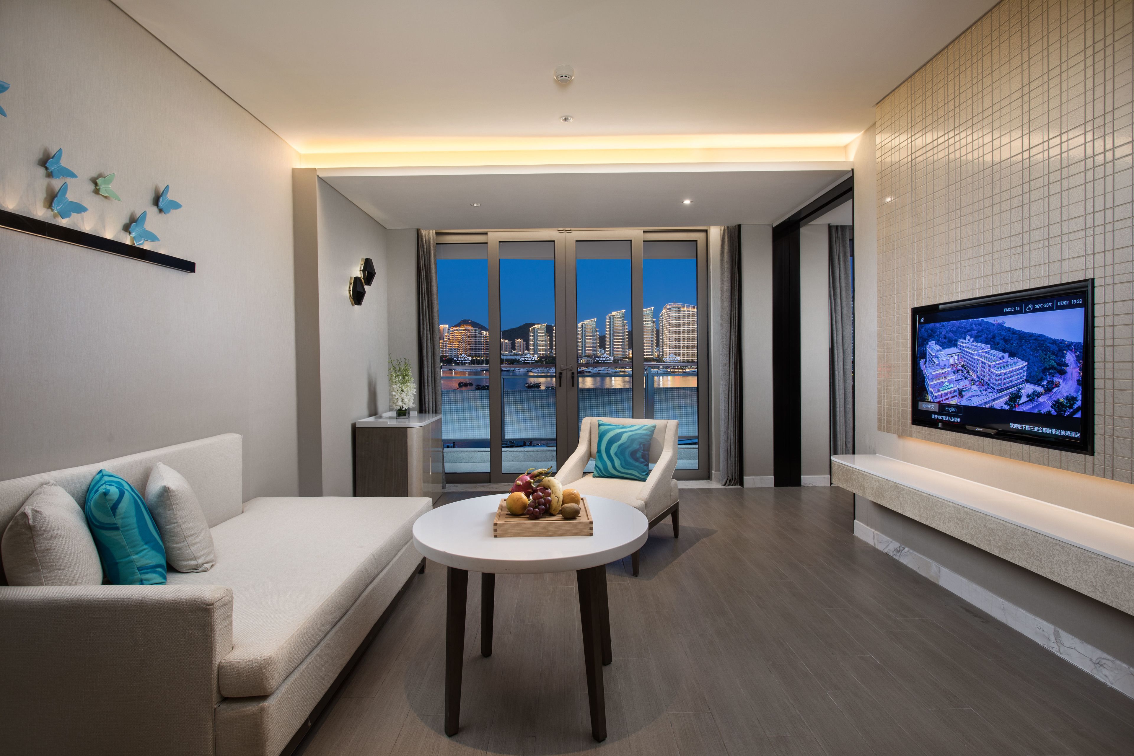 Ein modernes Wohnzimmer mit einer Couch, einem runden Tisch mit Früchten, einem Fernseher an der Wand und einer Stadtansicht durch große Fenster.