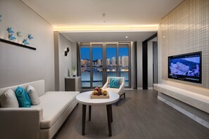 Un salon moderne avec un canapé, une table ronde sur laquelle sont disposés des fruits, un téléviseur fixé au mur et une vue sur la ville à travers de grandes fenêtres.