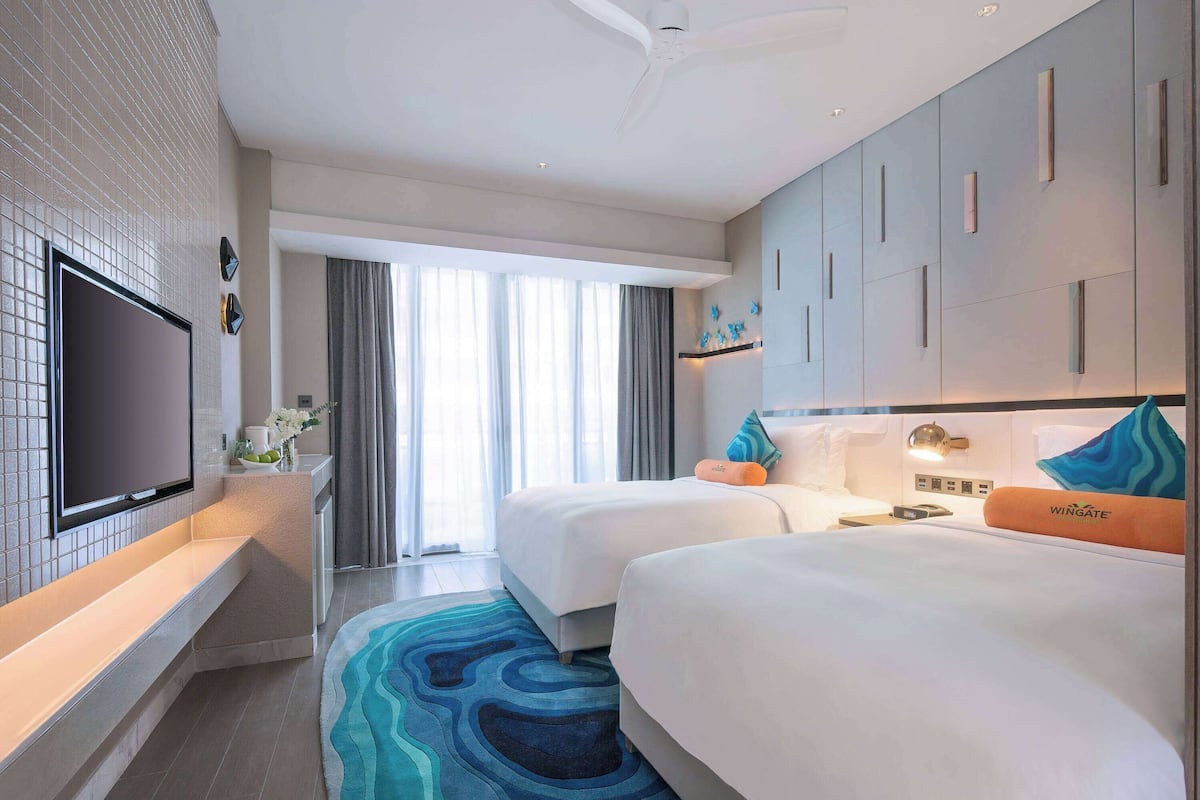 Een hotelkamer met een flatscreen televisie, twee bedden, een groot raam en een plafondventilator.