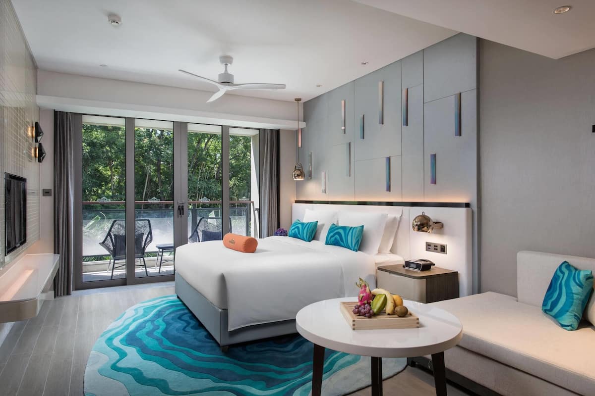 Een moderne hotelkamer met een groot bed, een bank, een tafeltje en een balkon met uitzicht op bomen.