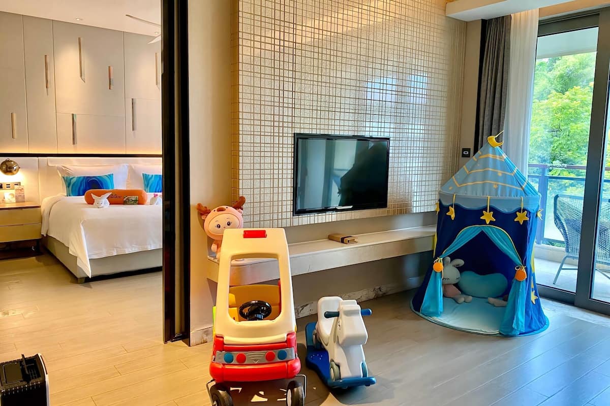 Een moderne hotelkamer met een bed, een televisie, een speelruimte met speelgoed en uitzicht op groen.