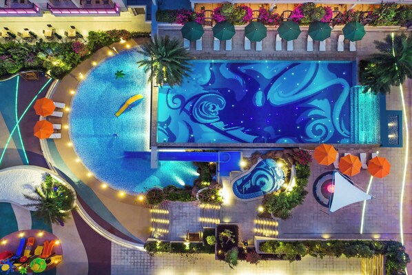 Vue aérienne d’un espace piscine d’hôtel, avec une fontaine centrale, des chaises longues disposées tout autour et des plantes décoratives.