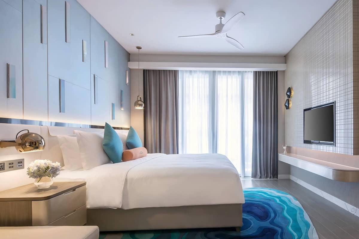 Een hotelkamer met een bed, een televisie, een plafondventilator en een groot raam met gordijnen.