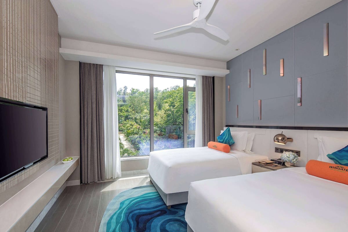 Hotelkamer met twee bedden, een televisie, een plafondventilator en een groot raam met uitzicht op groen.